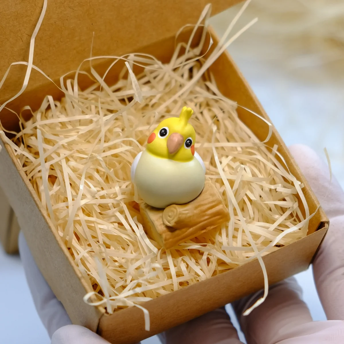 Whimsical handcrafted 3D cockatiel artisan keycap. It's vibrant, on a wooden stump with green grass and pink peonies.