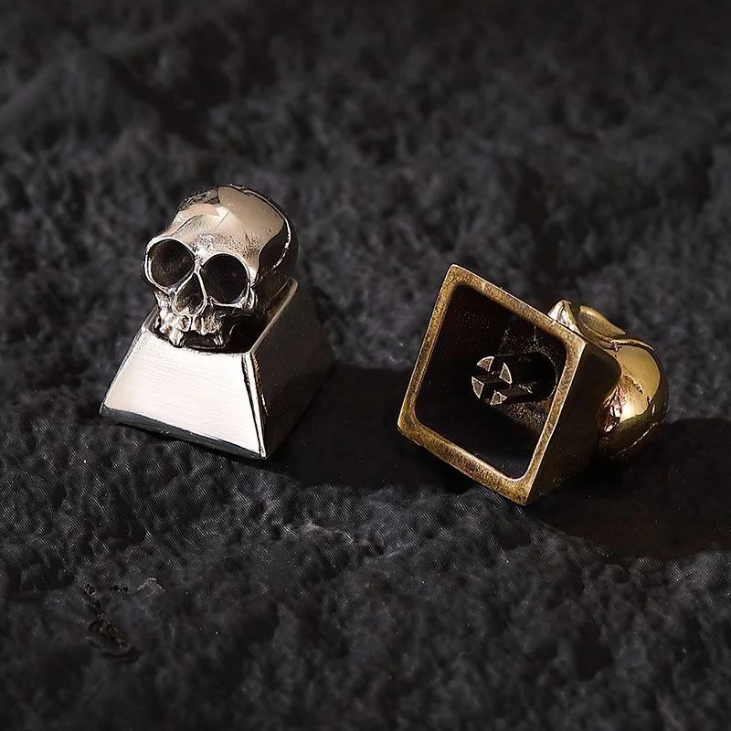 Two skull keycaps, one brass and one cupronickel, side-by-side on a dark surface.
